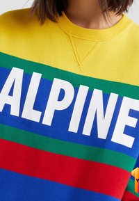Gul sweatshirts med djärv "ALPINE" text i vitt, med horisontella ränder i blått, grönt, rött och mörkblått över bröstet.