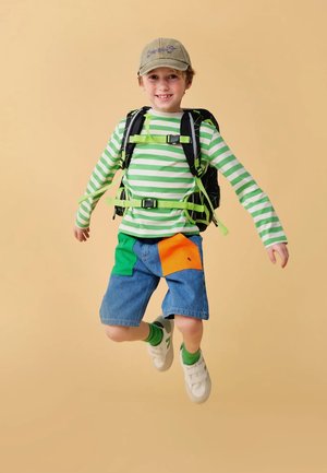 Grün-weiß gestreiftes Langarmhemd, Jeansshorts mit grünen und orangefarbenen Patches, schwarzer Rucksack und helle Sneakers.
