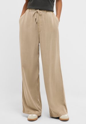 Person wearing beige wide-leg pants with drawstring waist and hands in pockets, paired with light-colored sneakers and a black top.