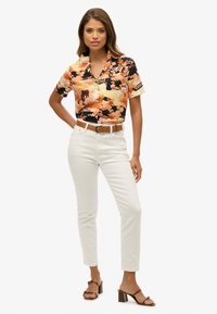Blusa negra de flores de manga corta con acentos en naranja y crema, combinada con pantalones blancos ajustados y sandalias de tacón marrones.