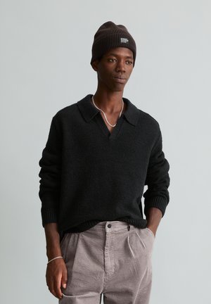 Homme portant un pull en maille noir, un bonnet marron, un collier chaîne argenté et un pantalon en velours côtelé marron clair, les mains dans les poches.