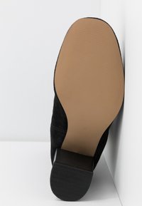 Semelle de botte en daim noir avec un avant lisse brun clair et un talon noir texturé, positionnée debout contre un fond blanc d'angle.