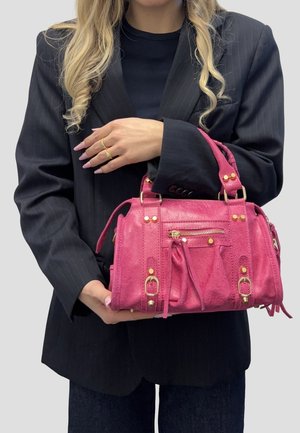 Donna con blazer scuro che tiene una borsa rosa testurizzata con accessori dorati e tasca frontale con cerniera, su sfondo neutro.