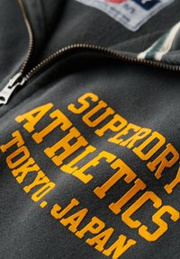 Sudadera gris con cremallera y letras en relieve amarillas; el texto dice "SUPERDRY ATHLETICS TOKYO, JAPÓN." La textura de la tela es suave y con relieve.