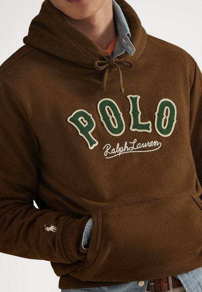 Καφέ φούτερ με κουκούλα, με πράσινα γράμματα "POLO" και τη λέξη "Ralph Lauren" γραμμένη, φορεμένο πάνω από ένα μπλε τζιν πουκάμισο, με τα χέρια στην μπροστινή τσέπη.