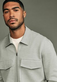 Light gray zip-up jacket with a spread collar, two front pockets, and a textured fabric. Worn over a cream sweater.