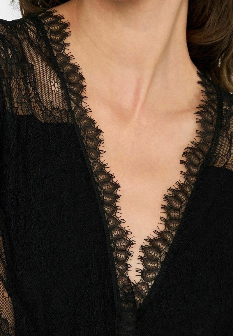 Gros plan d'une personne portant un vêtement en dentelle noire avec des bords festonnés détaillés le long d'un décolleté en V profond.