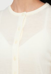 Κρεμ ζακέτα με κουμπιά, που διαθέτει στρογγυλή λαιμόκοψη, απαλό ύφασμα και τέσσερα κουμπιά ίδιου χρώματος κατά μήκος του μπροστινού μέρους.