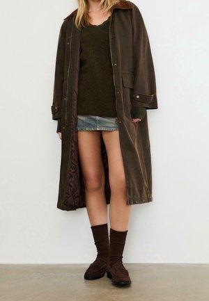 Mujer con abrigo largo marrón sobre suéter verde, minifalda de mezclilla, calcetines marrones y mocasines marrones, de pie contra una pared lisa.