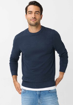 Mand iført en mørkeblå langærmet sweater over en hvid skjorte, stående med hænderne nær lommerne mod en ensfarvet lys baggrund.
