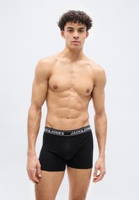 Νεαρός άνδρας με σγουρά μαλλιά, μυώδης, φορώντας μαύρα εσώρουχα boxer της Jack & Jones, στέκεται μπροστά σε απλό φωτεινό φόντο.