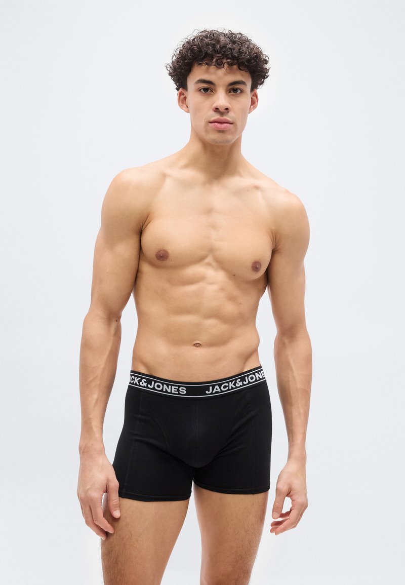 Νεαρός άνδρας με σγουρά μαλλιά, μυώδης, φορώντας μαύρα εσώρουχα boxer της Jack & Jones, στέκεται μπροστά σε απλό φωτεινό φόντο.