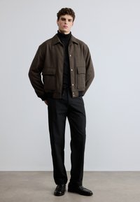 Hnedý vlnený kabát so zapínaním na gombíky a dvoma veľkými vreckami, nosený cez čierny rolák, v kombinácii s čiernymi nohavicami a topánkami.