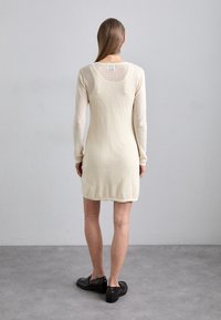 Abito in maglia beige chiaro a maniche lunghe, con texture in rete, scollatura rotonda sul retro e una silhouette leggermente informale. Sono indossate scarpe nere piatte.