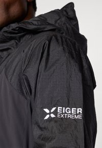 Zwarte jas met een gestructureerde afwerking, capuchon en geborduurd "EIGER EXTREME" logo op de mouw. Lichtgewicht en ontworpen voor gebruik in de buitenlucht.