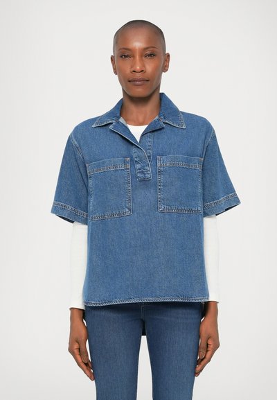 Chemise en denim à manches courtes bleu, avec un col pointu, deux poches avant et une coupe décontractée. Portée sur une chemise blanche à manches longues.
