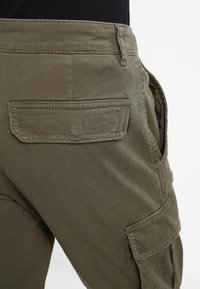 Vue arrière d’un pantalon cargo vert olive montrant les détails de la poche à rabat et de la poche latérale sur une personne portant un haut noir.