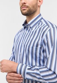 Camicia a righe in azzurro chiaro, blu navy e bianco. Presenta colletto con bottoni, taschino frontale e maniche lunghe con chiusura a bottoni. Texture del tessuto liscia.