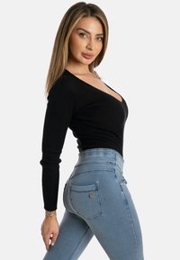 Pitkähiuksinen blondi nainen mustassa ribbineuletussa V-päällisessä ja vaaleansinisissä korkeavyötäröisissä farkuissa, poseeraa sivuttain yksiväristä taustaa vasten.