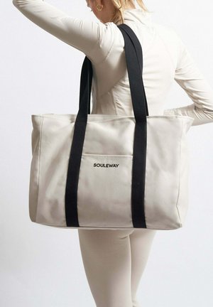 Beige Tragetasche mit schwarzen Trägern, ausgestattet mit einer Fronttasche und dem Logo 'SOULEWAY'. Rechteckige Form mit glatter Textur.