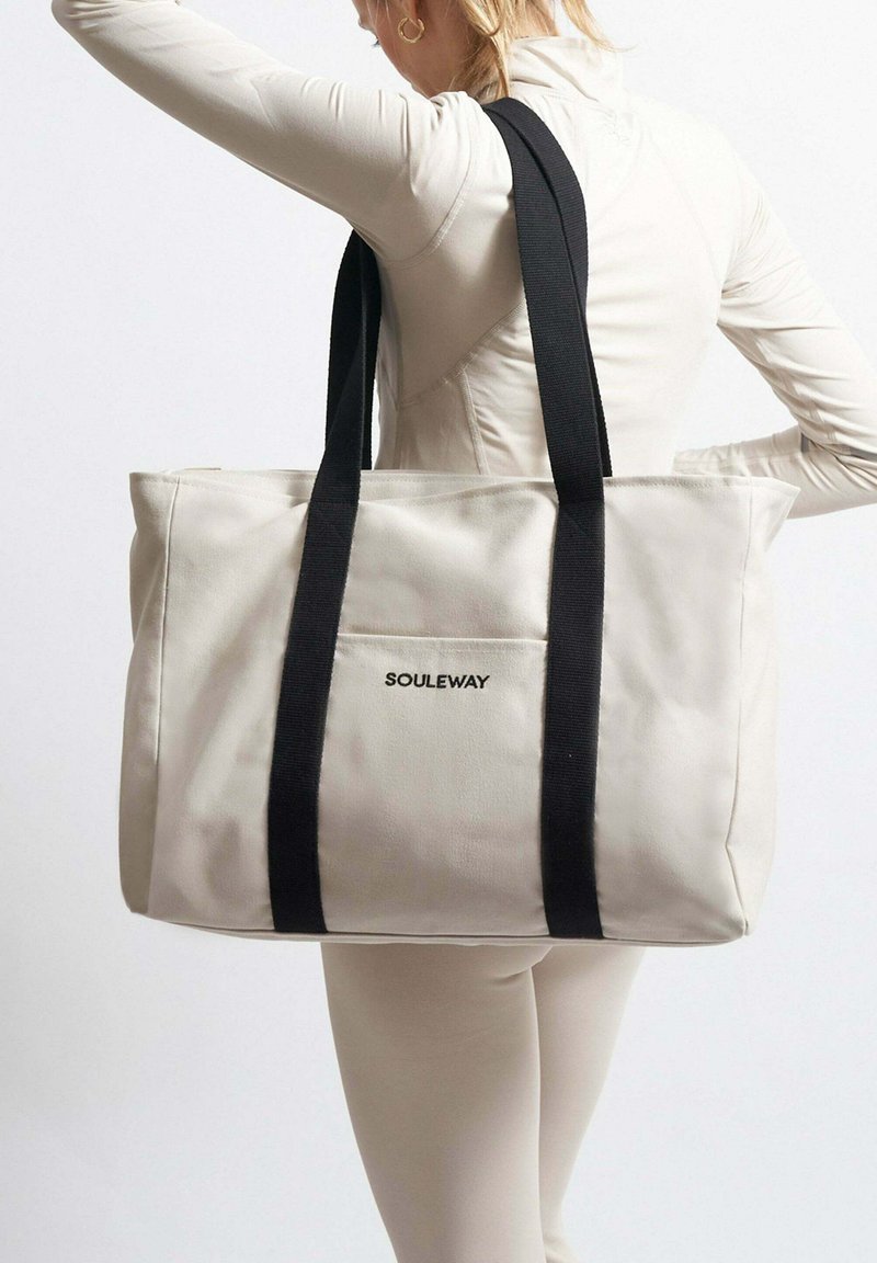 Béžová taška přes rameno s černými popruhy, se zadní kapsou a logem 'SOULEWAY'. Obdélníkový tvar s hladkým povrchem.