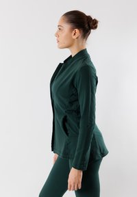 Giacca verde scuro con un design aderente, tasche laterali e colletto alto. Tessuto liscio con un leggero lucido e cuciture sartoriali.