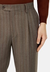 Pantaloni di lana grigi con strisce verticali marroni, dotati di davanti piatto, passanti per cintura e chiusura con bottone. Mano appoggiata sulla vita.