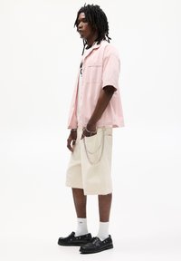 Camicetta rosa chiaro a maniche corte abbinata a pantaloni beige. L'outfit presenta un accessorio in catena argentata e scarpe nere, con calzini bianchi.