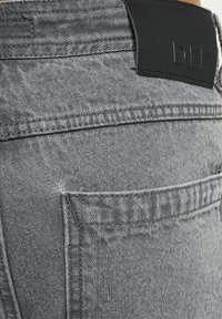 Harmaat farkut, joissa on teksuroidun pinnan, varustettuna mustalla nahkavyöpaikalla, jossa on kohokuvioidut kirjaimet, sekä takataskulla.