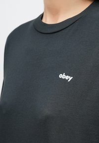 Dunkelgraues T-Shirt mit rundem Ausschnitt und kurzen Ärmeln. Auf der Vorderseite prangt ein weißes "obey"-Logo, gefertigt aus einer weichen Baumwollmischung.