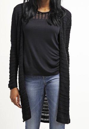 Frau trägt eine lange, offene schwarze Strickjacke über einem schwarzen Oberteil mit Mesh-Details, kombiniert mit hellblauen Jeans.
