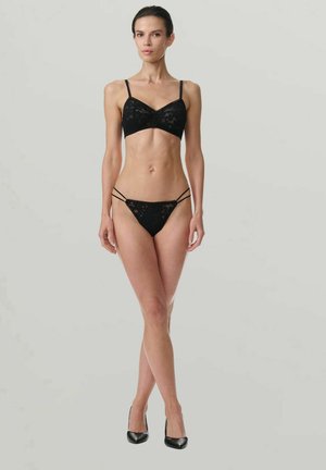 Vrouw die tegen een effen achtergrond staat, gekleed in een zwarte kanten bralette, bijpassend strappy kanten ondergoed en zwarte hoge hakken.