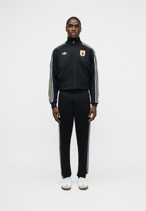 Άνδρας που φοράει μαύρη φόρμα Adidas με λευκές ρίγες και αθλητικό έμβλημα, στέκεται μπροστά σε απλό λευκό φόντο.