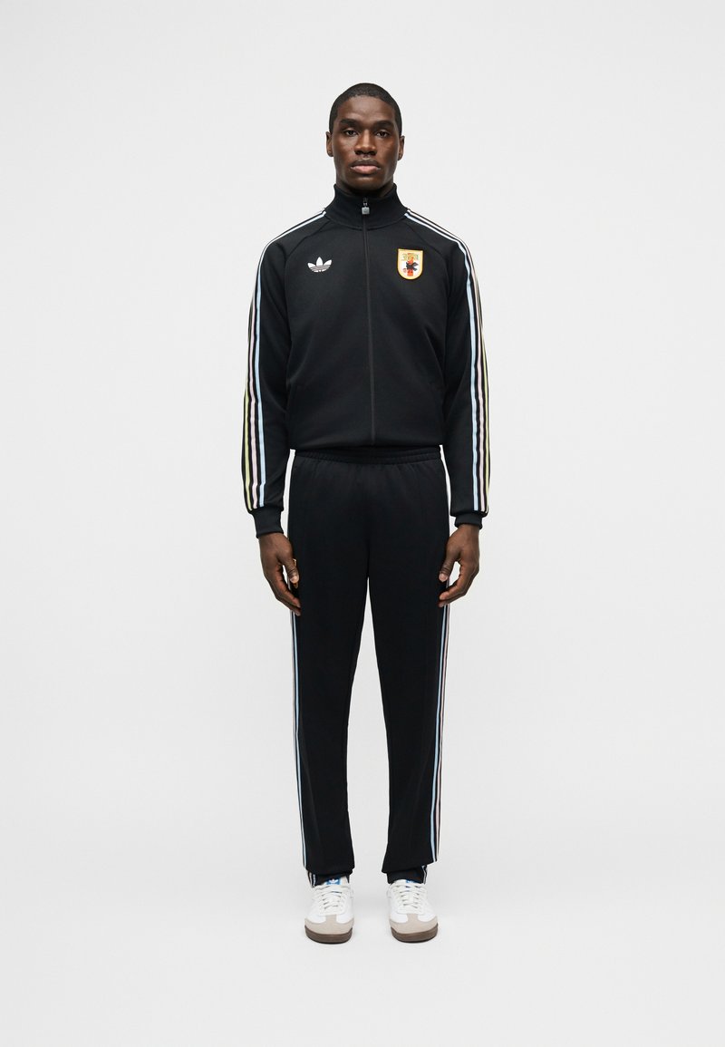 Mees mustas Adidas dressipluusis valgete triipude ja spordiembleemiga, kes seisab ühevärvilise valge tausta ees.