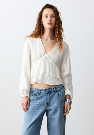 Jeune femme aux longs cheveux bouclés portant un chemisier blanc à manches longues avec un détail noué à l'avant et un jean bleu clair, sur un fond uni.