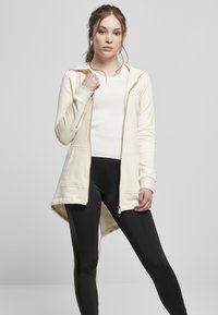 Sudadera larga color crema con cremallera en el frente, dos bolsillos delanteros y un dobladillo posterior más largo, combinada con leggings negros. Textura suave.