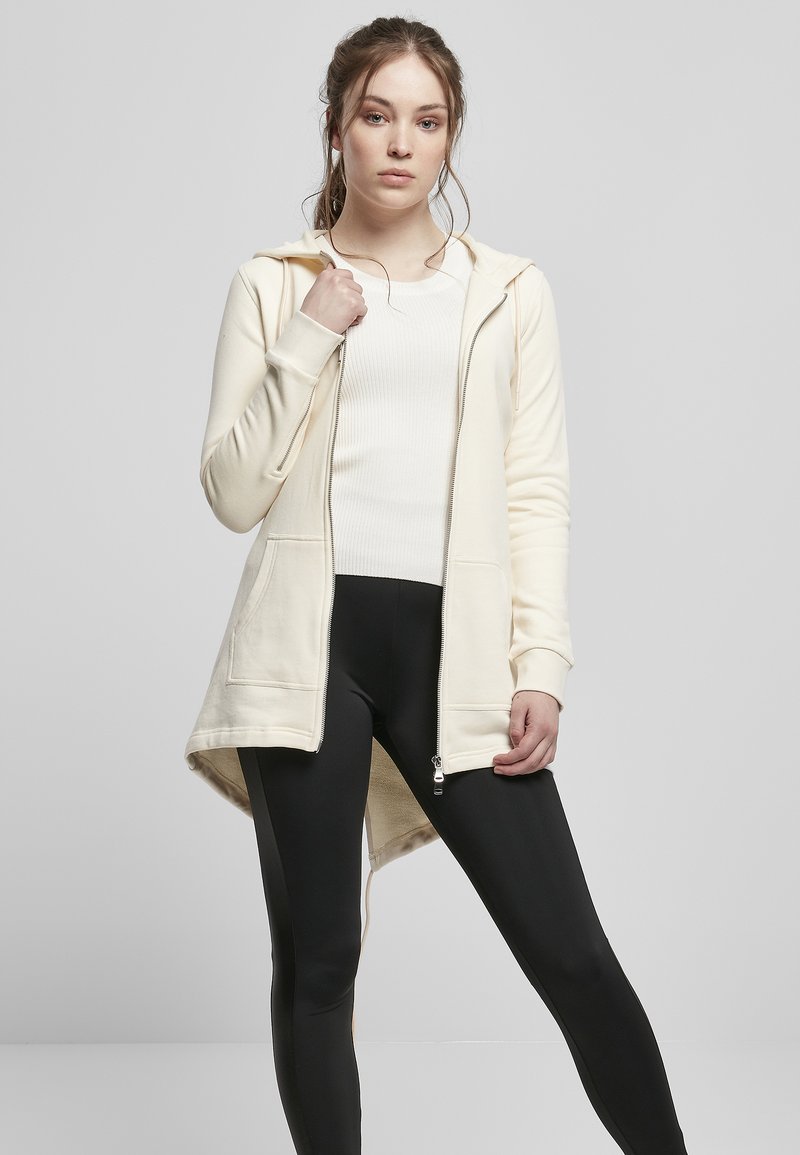 Sudadera larga color crema con cremallera en el frente, dos bolsillos delanteros y un dobladillo posterior más largo, combinada con leggings negros. Textura suave.