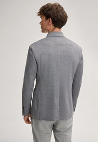Grijze herringbone blazer met een gespreide kraag, een gladde textuur, twee zijzakken en knoopmanchetten, gedragen over lichtgekleurde broeken.
