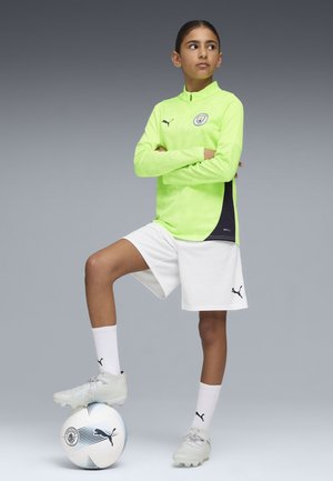 Maillot à manches longues vert fluo avec des accents noirs sur les côtés, shorts blancs, chaussettes de foot blanches et chaussures grises claires, posant avec un ballon de soccer.