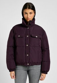 Giacca a vento di colore viola scuro, con colletto alto, bottoni a pressione e due tasche frontali. Il materiale è morbido e trapuntato, con polsini elastici.
