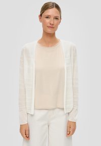Leichter, offener Cardigan in Weiß mit strukturierten Streifen, kombiniert mit einem transparenten beigen Top. Das Model trägt weiße Hosen.