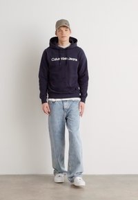 Tengerészkék kapucnis pulóver "Calvin Klein Jeans" logóval, világoskék, bő szárú farmernadrággal és fehér sportcipővel párosítva. A modellen szürke sapka látható.