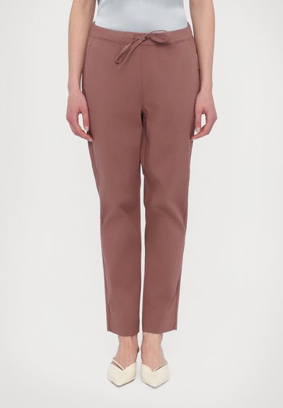 Pantalons cintrés de couleur rouille avec une ceinture à cordon. Le matériau a l'air doux, avec une texture lisse. Portés avec des chaussures pointues de couleur claire.