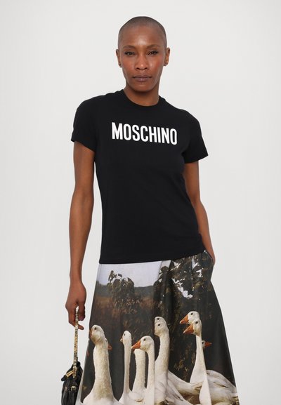 Melna kokvilnas t-kreklā ar baltu "MOSCHINO" logotipu, apvienota ar svārkiem, kuru dizainā ir fotogrāfijas druka ar baltiem gailītiem dabiskā fona. 