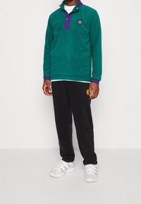 Grön fleecetröja med lila krage och knappar, svarta träningsbyxor med en cirkulär logotyp, bärs med vita träningsskor.