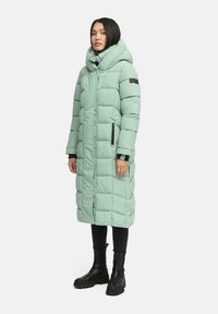 Lange, mintgroene pufferjas met een capuchon, gewatteerd ontwerp, zijzakken en zwarte accenten. Gdragend met zwarte skinny jeans en stevige zwarte laarzen.
