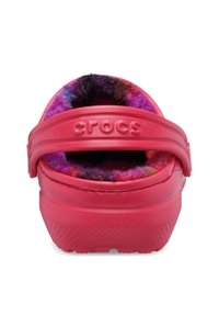 Różowe Crocs z teksturowaną podeszwą i puszystym purpurowo-czarnym wnętrzem. Posiadają szeroki pasek na pięcie i gładki materiał zewnętrzny.