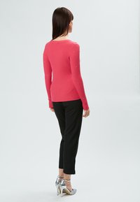 Taillierter, gerippter Pullover in leuchtendem Pink mit langen Ärmeln und dekorativen Nieten, kombiniert mit maßgeschneiderten schwarzen Hosen und silbernen High-Heels.