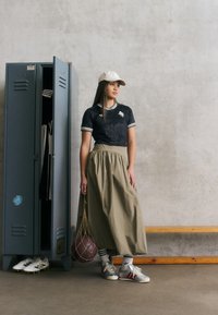 Crna majica kratkih rukava s prugastim kragnom, bež duga suknja, bijela kapa i sivi tenisici; drži smeđi lopti u mrežastoj torbi.