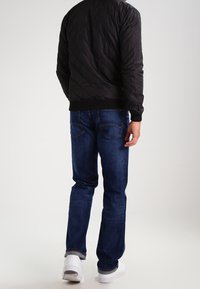 Mann trägt eine dunkle Steppjacke, blaue Jeans und weiße Sneakers und steht mit dem Rücken zu einem schlichten Hintergrund.